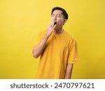 A young man in a yellow shirt yawns with his hand over his mouth against a bright yellow background.