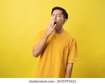 A young man in a yellow shirt yawns with his hand over his mouth against a bright yellow background. - Powered by Shutterstock