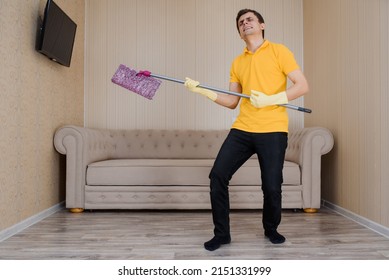 Young Man In A Yellow T-shirt And Yellow Gloves Is Cleaning The Wood Flooring Using Floor Mop And Having Fun Plays Guitar