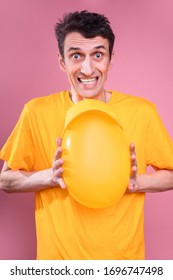 Young Man In Yelow Shirt Squeeze Balloon With Both Hands. Trying Hard. Isolated Over Pink Background