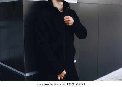 Young Man Zipping Up His Hoodie. Man Dressing Black Sport Clothes. Grey Urban Background. 