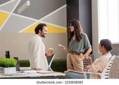 young manager holding paper cup near smiling colleague with smartphone and multiracial businesswoman sitting in office - Powered by Shutterstock