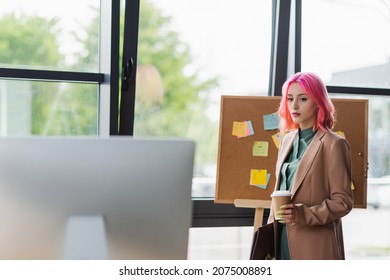 young manager with pink hair and piercing holding paper cup and looking at computer monitor
