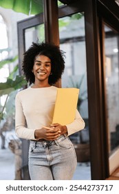 Young manager is smiling while holding a yellow folder and a pen, standing in a modern coworking space, embodying diversity and social inclusion in the workplace