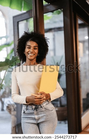 Young manager is smiling while holding a yellow folder and a pen, standing in a modern coworking space, embodying diversity and social inclusion in the workplace
