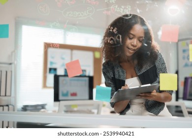 Young manager taking notes on clipboard while analyzing project plan and data on sticky notes in modern office