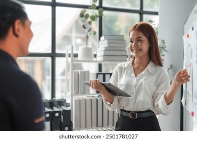 Young manager woman is holding a digital tablet and presenting a business plan on a whiteboard to her colleague - Powered by Shutterstock