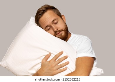 Young manly bearded man with closed eyes hugging soft and comfortable white pillow with delightful smile, sleeping like baby, enjoying sweet dreams, feeling relaxed, isolated over gray studio wall - Powered by Shutterstock