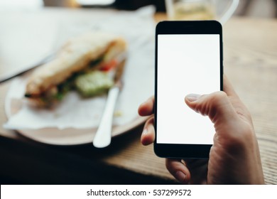 Young Man's Hand Holding Mobile Phone. He Is Sitting In Restaurant Or Cafe. He Is Scrolling And Reading Some Stuff. Place For Your Advertisment Or Logo. Back View. Horizontal Position Of The Phone