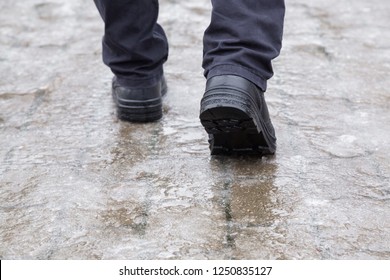 Young Man's Legs In Black Leather Boots Walking On Sidewalk In Wet, Warm Winter Day. Pavement Covered With Slippery Ice. Closeup. Back View.