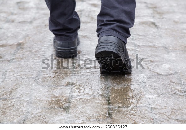 Young man\'s legs in black leather boots walking on\
sidewalk in wet, warm winter day. Pavement covered with slippery\
ice. Closeup. Back view.