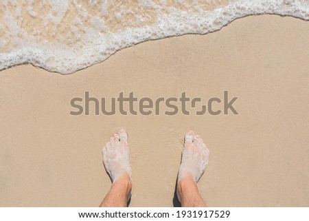 The young man's legs stand on the beach sand and are washed by blue sea water, top view.