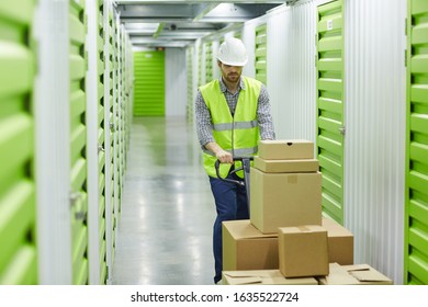 Young manual worker in work helmet carrying cardboard boxes on cart while working in warehouse - Shutterstock ID 1635522724