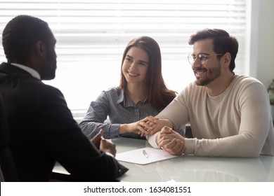 Young married couple and African insurance agent communicating seated in office room, worker explain features of various policies, contacting with potential customers ready to sign agreement concept