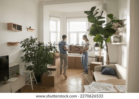 Young married couple carrying boxes into rented flat, cozy room with houseplants, moving into new home after renovation, buying apartment, enjoying relocation. Full length shot
