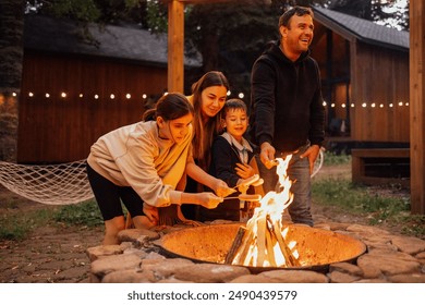 A young married couple with children roasts marshmallows on the fire. A man with his wife, son and daughter are having fun in the backyard of the house. A happy family outdoors near a campfire - Powered by Shutterstock