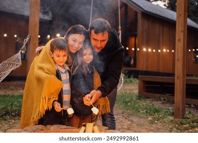 A young married couple with children roasts marshmallows on the fire. A man with his wife, son and daughter are having fun in the backyard of the house. A happy family outdoors near a campfire - Powered by Shutterstock