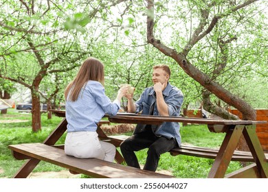 young married couple in denim clothes sitting in the garden at a wooden table and drinking coffee on a date in the summer, girl and guy having breakfast in an outdoor cafe and chatting - Powered by Shutterstock