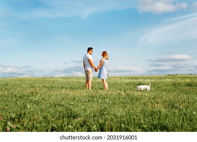 Young Married Couple With Dog On Walk In Green Field, Hold Hands And Look At Each Other Against Blue Sky Outdoors. Family Spending Time Together, Leisure.