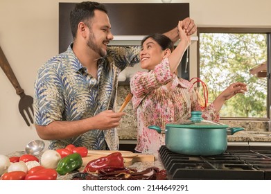 A Young Married Couple Happily Dancing Caribbean Music In The Kitchen While Preparing Food.