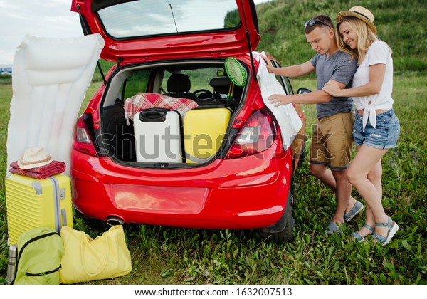 Young married couple\
looking at a map of the area, camping in nature. Red car loaded\
with Luggage for travel