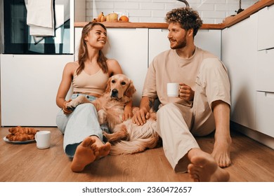 A young married couple in love sitting on the floor in the kitchen drinking coffee in the morning with their beloved dog, romantically spending time together. - Powered by Shutterstock