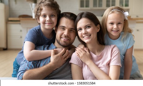 Young Married Couple Loving Parents Their Little Adorable Kids Daughter And Son Seated Together On Couch Embracing Pose Looking At Camera, Concept Of Happy Wealthy Family Enjoy Time At New Modern Home