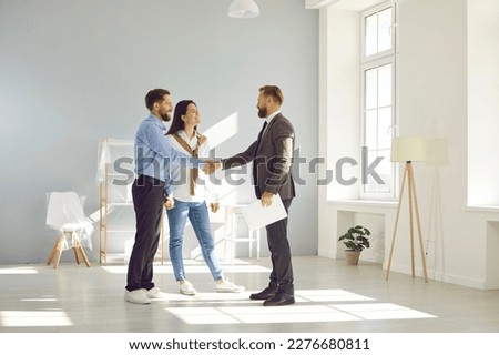 Young married couple make deal, buy new house and exchange handshake with relator. Happy man and woman standing in modern light spacious light living room and shaking hands with real estate agent