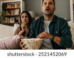 Young married couple, man and woman, sitting at home on sofa, eating popcorn and watching scary horror movie or dramatic sports games action or drama film. Simple living concept, weekend activities