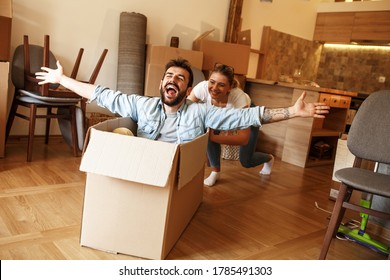 Young married couple moving into a new home.Man sitting in cardboard box while woman pushes him all over the room.Real estate funny concept 
