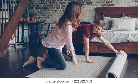 Young Married Couple Is Rolling Out Beautiful New Carpet Ion Floor N Bedroom At Home Then Enjoying It. House Interior, Married Life And Happy People Concept.