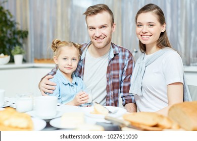 Young married couple and their sweet daughter looking at camera while sitting by served table in the kitchen