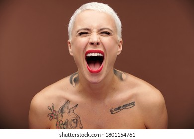 Young Masculine Female With Short Blond Hair, Red Lipstick On Lips And Tattoos On Chest And Neck Shouting And Expressing Aggression