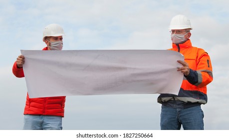 Young And Mature Engineers Or Workers Discussing A Construction Plan With Face Mask And Social Distancing - Focus On The Faces