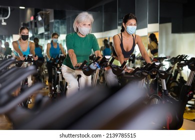 Young And Mature Female In Face Masks Warming Up On Bikes In Cycling Class At Gym