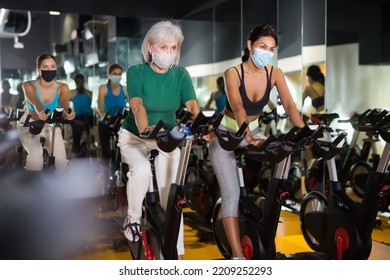 Young And Mature Female In Face Masks Warming Up On Bikes In Cycling Class At Gym