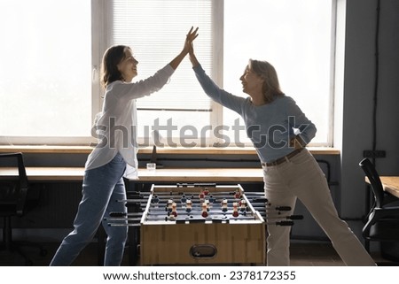 Young and mature women, two office employees playing table football, give high five, celebrate victory feel happy, enjoy lunch break together at workplace. Friendship, fun at workplace, companionship