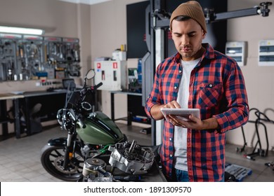 young mechanic in beanie and plaid shirt using digital tablet near spare parts and motorcycle on blurred background