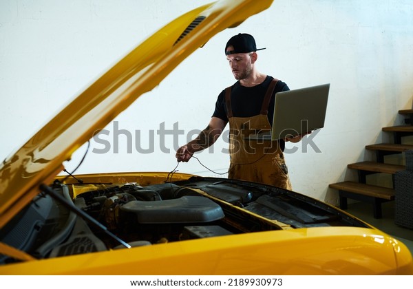 Young mechanic with laptop installing wires\
into engine of car while carrying out technical diagnostics of\
motor after repairment