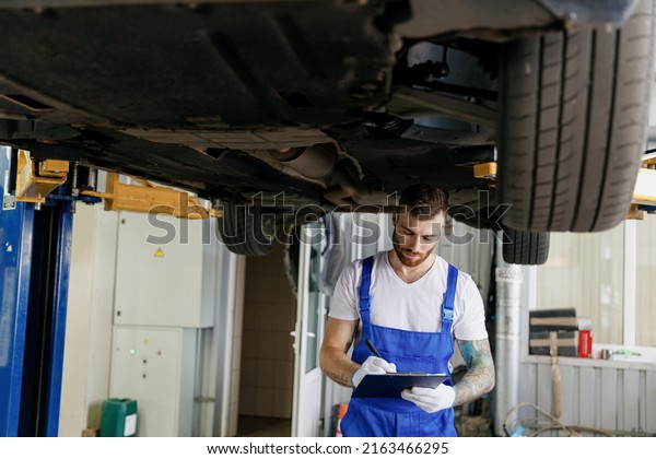 Young mechanic man in denim overalls use hold\
clipboard document writing estimate outlay stand near car lift\
check technical condition work in vehicle repair shop workshop\
indoor. Tattoo translate\
fun