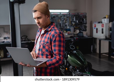 young mechanic in plaid shirt and beanie using laptop in workshop near motorcycle on blurred background - Powered by Shutterstock