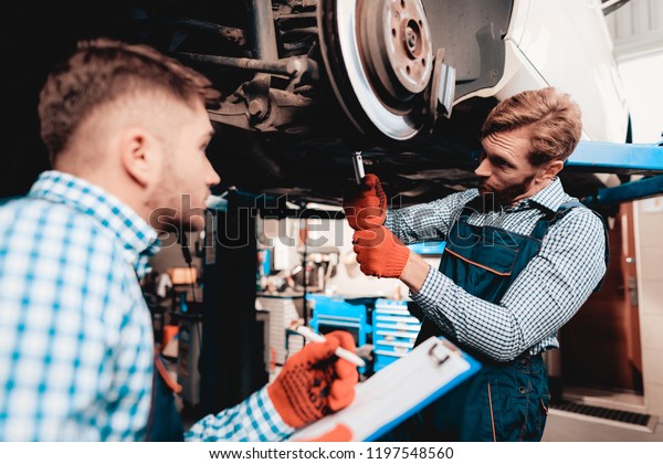 Young Mechanic Repairs Automotive Hub In
Garage. Professional Uniform. Service Station Concept. Confident
Engineer Stare. Detail Repairing. Under The Vehicle. Automobile
Diagnostic.