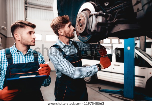 Young Mechanic Repairs Automotive Hub In\
Garage. Professional Uniform. Service Station Concept. Confident\
Engineer Stare. Detail Repairing. Under The Vehicle. Automobile\
Diagnostic.