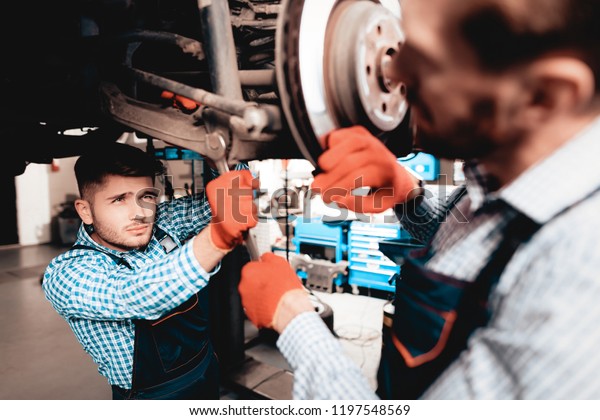 Young Mechanic Repairs Automotive Hub In Garage.\
Professional Uniform. Service Station Concept. Confident Engineer\
Stare. Detail Repairing. Under The Vehicle. Automobile Diagnostic.\
Join Forces.