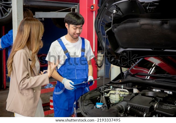 Young mechanic talking to customer asian woman\
about car with repairing in garage, auto service, technician man\
explaining problem of vehicle and look clipboard with client,\
transportation concept.