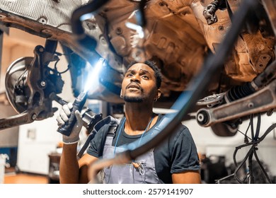 Young mechanic using flashlight, inspecting underside of car in a professional workshop, focused on maintenance and repair