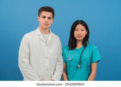 Young medical professionals in white coat and scrubs posing together against blue background; confident doctors or healthcare team ready for work; dedicated nurses and physicians representing modern