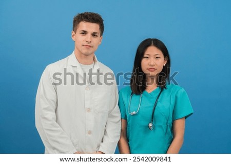 Young medical professionals in white coat and scrubs posing together against blue background; confident doctors or healthcare team ready for work; dedicated nurses and physicians representing modern