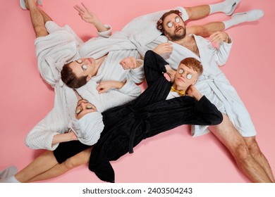 Young men, friends with cucumber eye mask lying on floor in bathrobes against pink background. Holidays preparation, friends supporting groom. Concept of leisure, fun, bachelor party, friendship, spa - Powered by Shutterstock