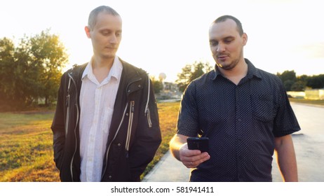Young men have a serious talk while walking in the city during beautiful sunset with lense flare effects. Looking at mobile phone businessman showing something about new project